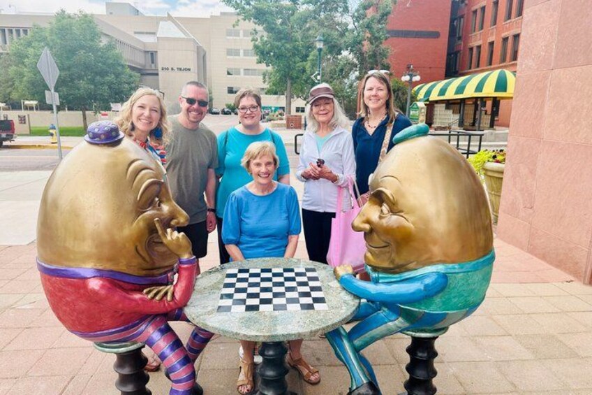 A group of people standing around a chess table with two large, colorful, anthropomorphic figures on either side. 4