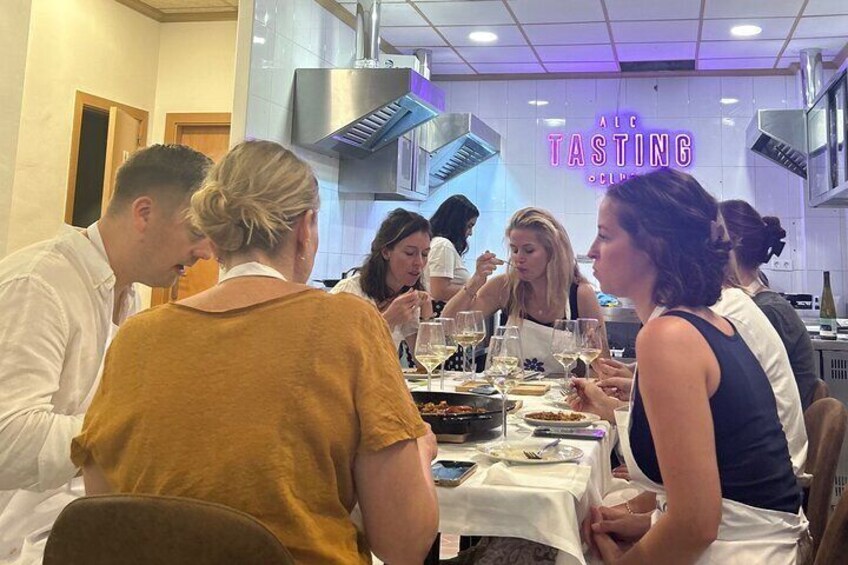 En grupp människor äter middag på en restaurang med en neonreklamskylt som visar texten "ALC TASTING CLUB". 2