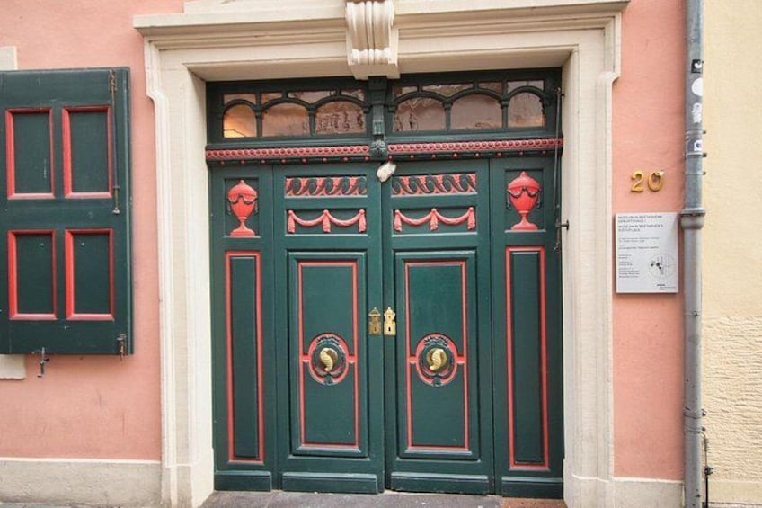 A double green door with red trim and a decorative pattern, flanked by a pink wall and a number plaque. 2
