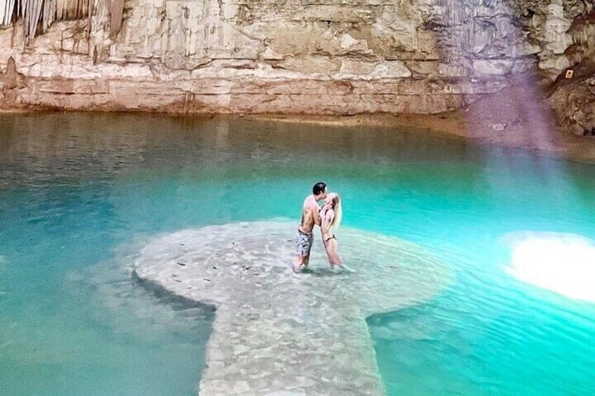 A person standing on a circular stone platform in a clear blue pool with a rocky cliff in the background. 3