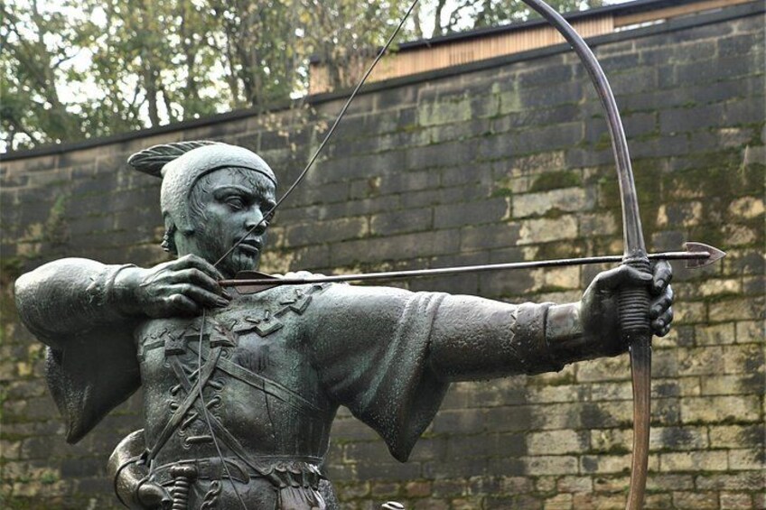 A bronze statue of a figure with a bow, wearing a feathered headdress, set against a stone wall. 3