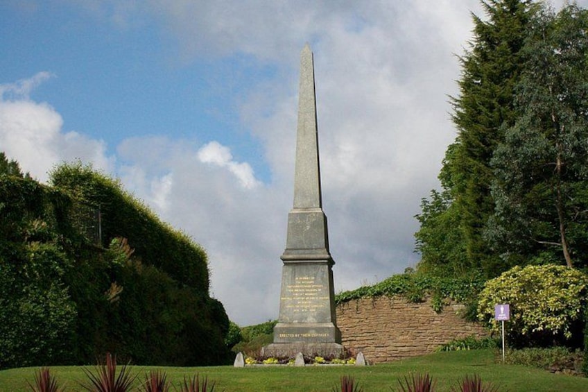 A tall, narrow monument with inscriptions, surrounded by well-maintained grass and hedges, with trees in the background. 5