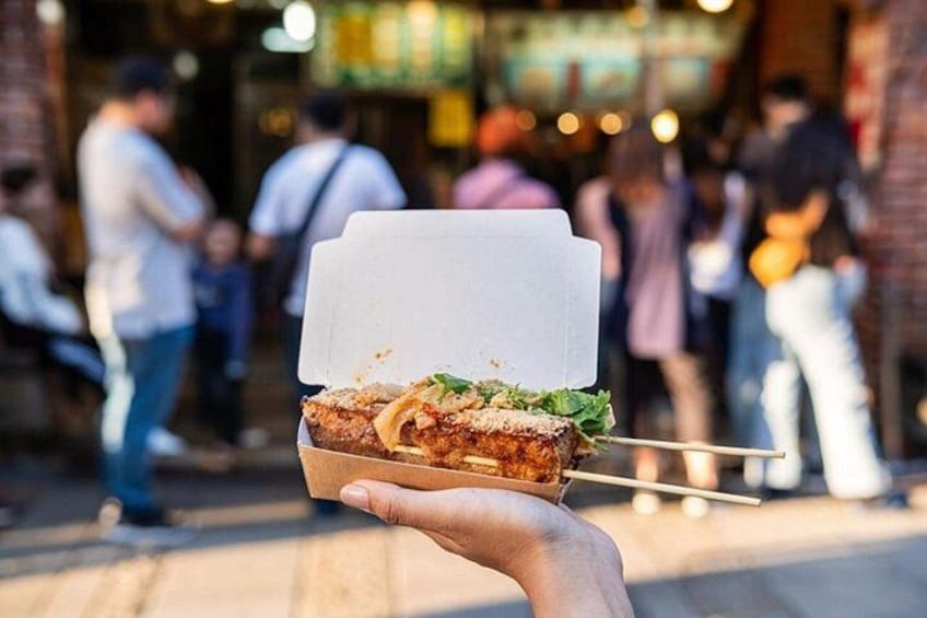 Una mano sosteniendo un alimento ensartado en un palillo con un envoltorio de papel blanco, sobre un fondo de calle concurrida con gente y edificios. 2