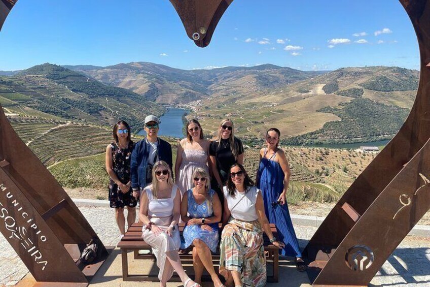 A group of people posing in a heart-shaped structure with a scenic view of hills and a lake in the background. 5
