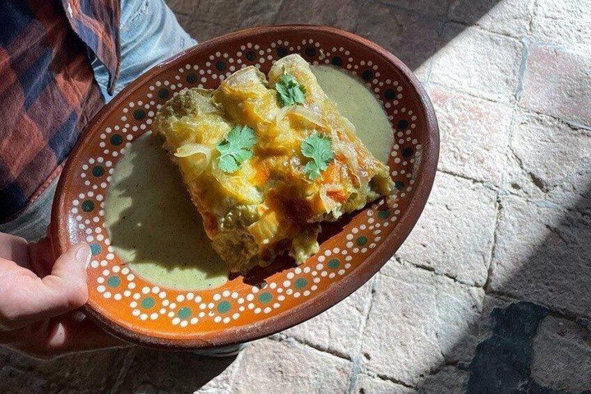Een persoon houdt een traditioneel kleibordje vast met een gerecht erop, mogelijk een gevuld groente- of vleesgerecht, versierd met een groen kruid. 2
