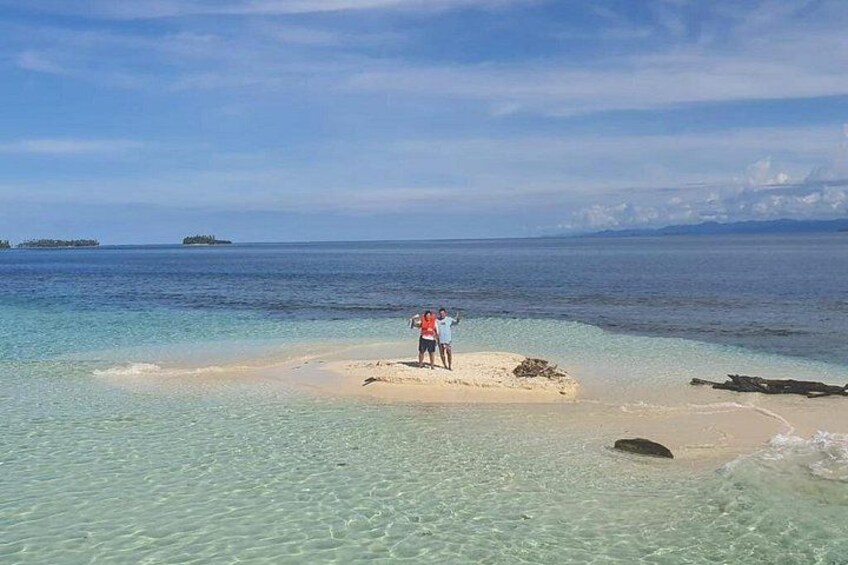 A person standing on a small sandy island in clear turquoise water, with distant islands visible on the horizon. 3
