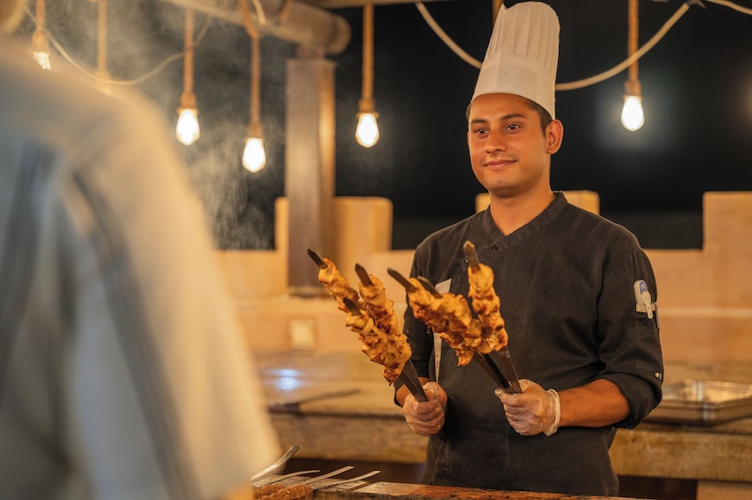 Un chef sostiene brochetas de comida, con una persona desenfocada al frente. 5