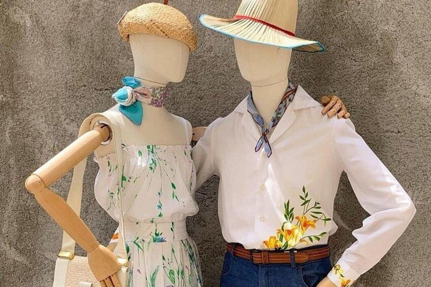 Un mannequin vêtu d’un chemisier blanc et d’une robe à motifs floraux, se tenant à côté d’un autre mannequin portant une chemise blanche et un jean bleu, tous deux placés devant un mur à texture. 1