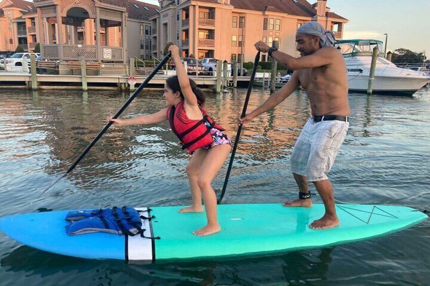 A person paddleboarding on a turquoise board with a life jacket, assisted by another person. 3
