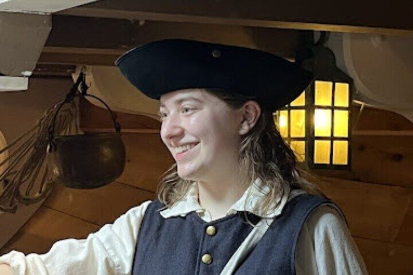 A person in historical attire, including a dark hat and a vest, in a wooden interior with a hanging pot and a window with warm light. 2