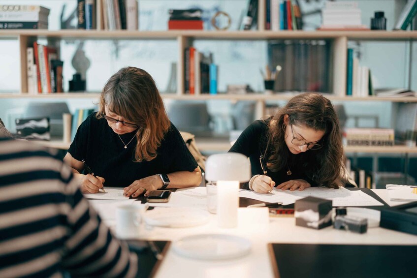 Two individuals writing at a table with books and a lamp in the background. 1