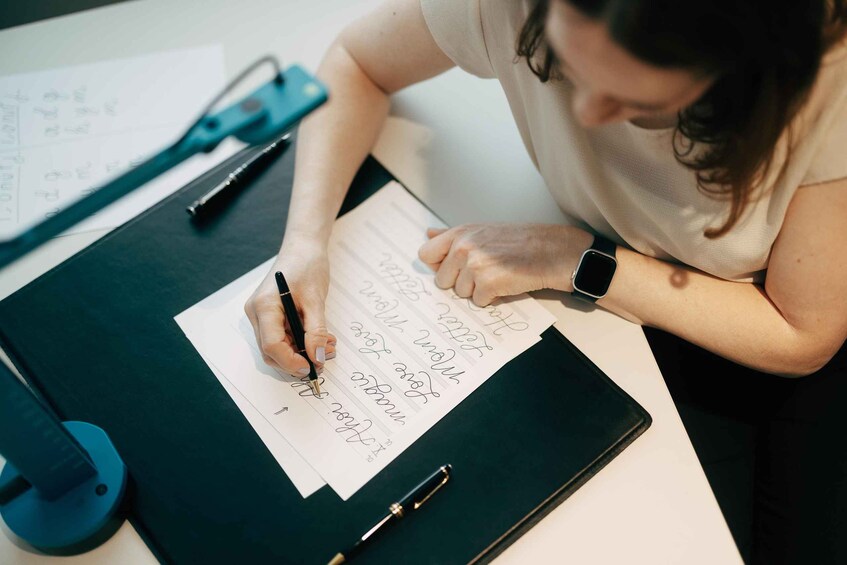 A person writing on a piece of paper with a pen, a notebook, and a smartphone on a desk. 4