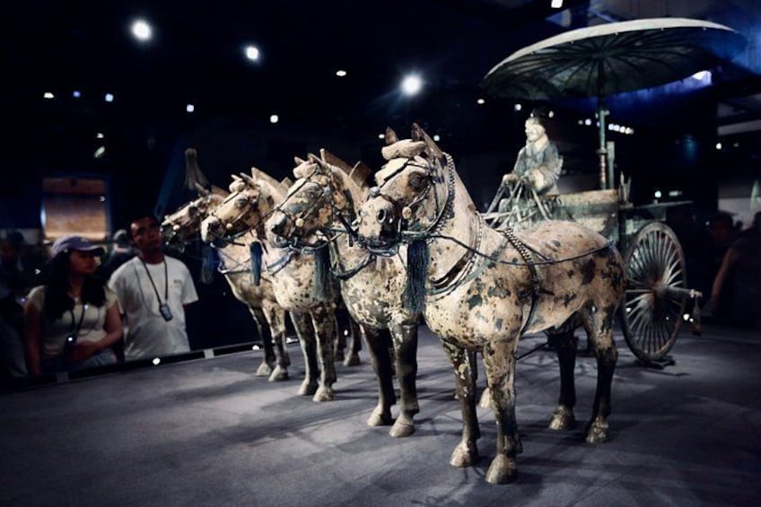 Een historisch overzicht van wagens met paarden, een persoon zittend in een wagen, en een grote wielstructuur op de achtergrond. 1