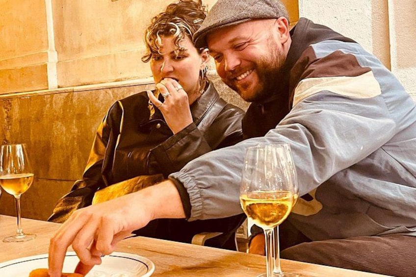 Två personer sitter vid ett bord med vinglas, och en av dem sträcker sig efter en tallrik. 1