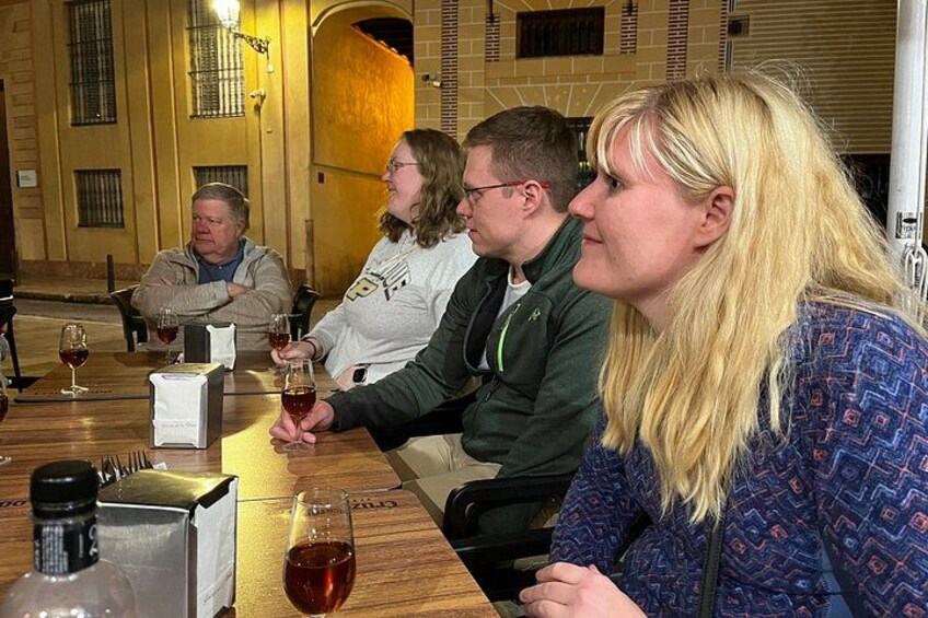 En gruppe mennesker sidder ved et bord med vinglas, en flaske og en æske i et rum med murstensvægge og et vindue. 2