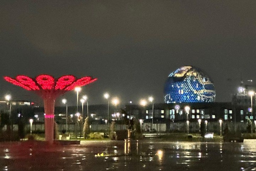 Una escultura iluminada con pétalos rojos, un gran globo luminoso y un paisaje urbano de fondo. 3