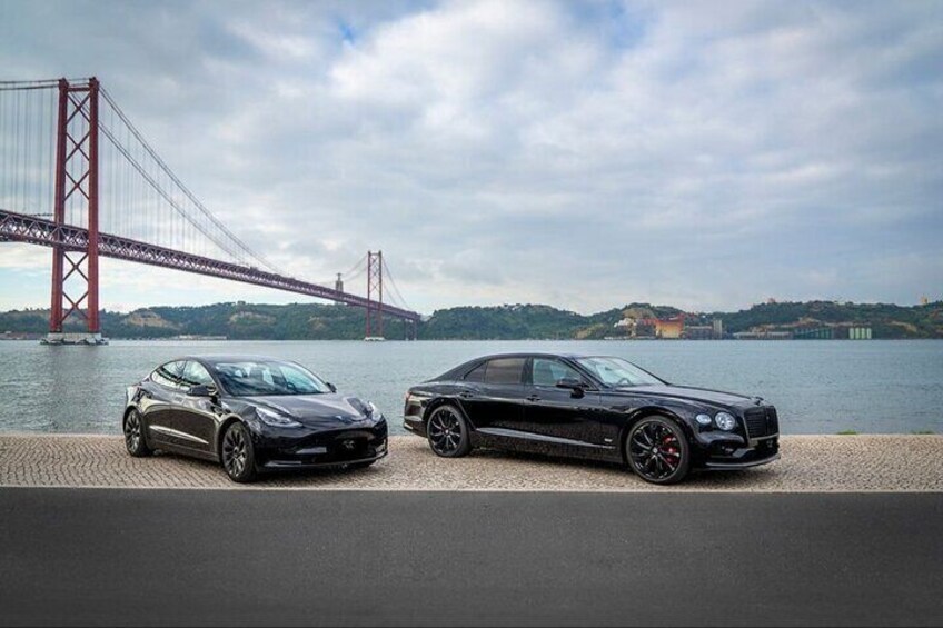 Two cars parked by a waterfront with a bridge in the background. 1