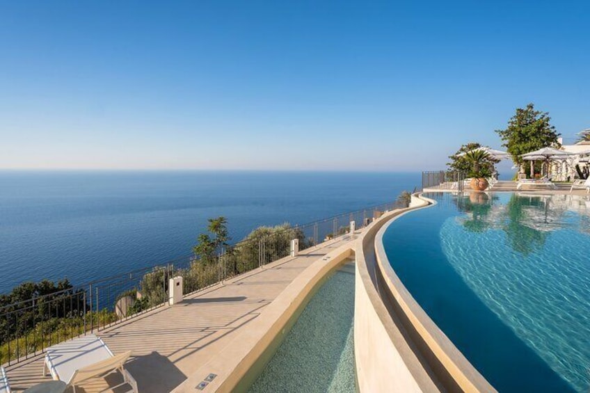 Una piscina infinita curva con vistas al océano, con sillones de descanso y una zona sombreada. 3