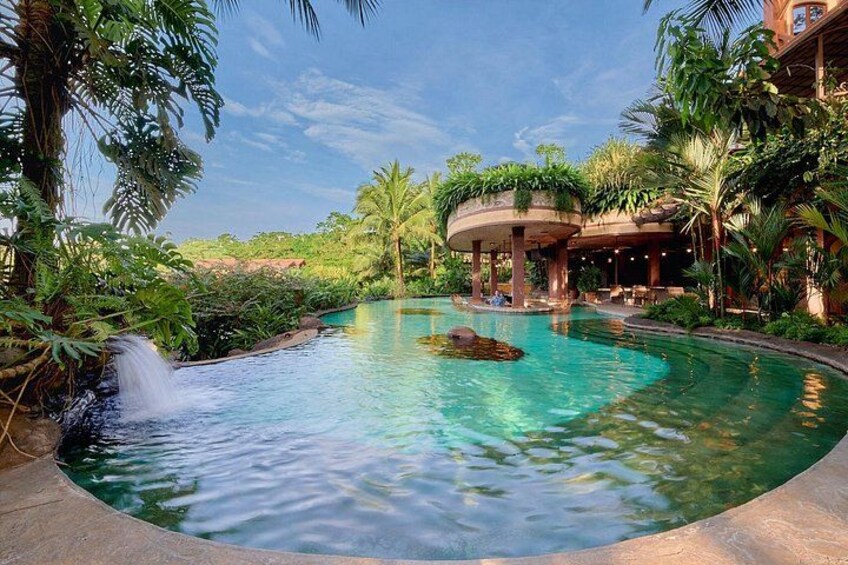 A pool with a waterfall, surrounded by lush greenery and a covered area with plants. 2