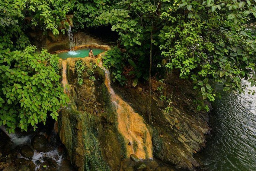 A natural hot spring surrounded by lush greenery and a rocky waterfall. 4