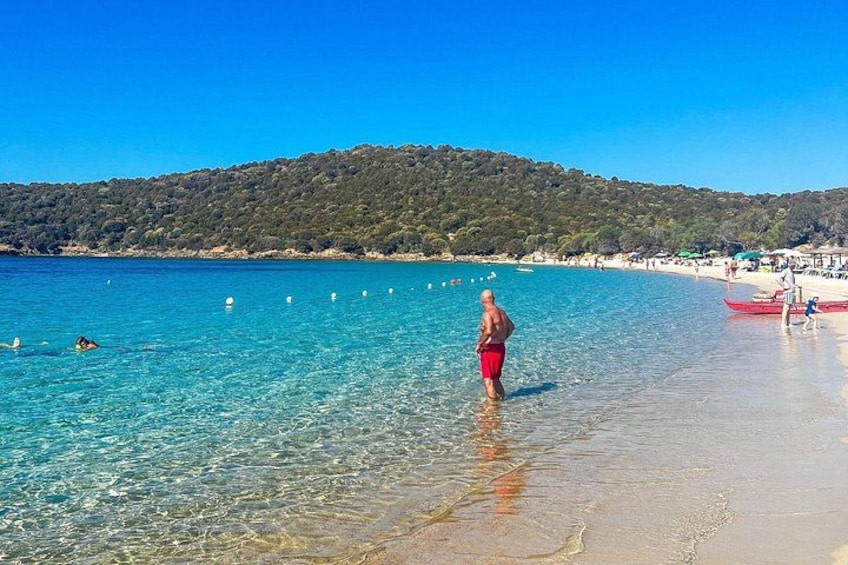 A beach with clear turquoise water, a person standing in shallow water, and a boat on the shore. 2