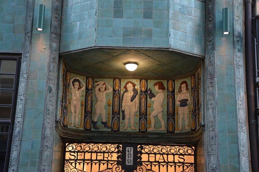 La façade d’un immeuble est ornée d’une mosaïque représentant des figures humaines. On y voit une enseigne indiquant « SELSER », et en dessous, une autre enseigne portant l’inscription « CATHS ». 3