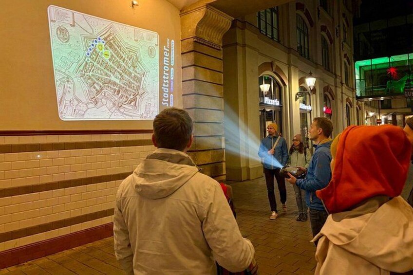 Une projection d’une carte sur un mur, des personnes debout qui la regardent, et un bâtiment aux fenêtres en arc de cercle en arrière-plan. 1