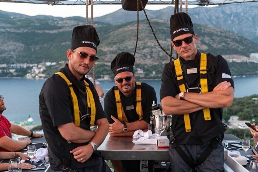 Three men wearing chef hats and yellow suspenders, standing at a table with a lake and mountains in the background. 4