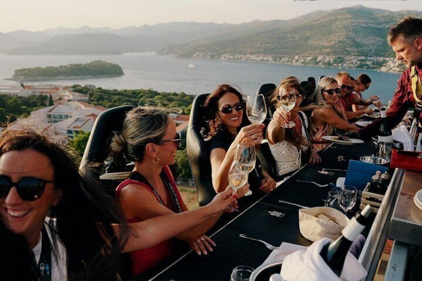 A group of people toasting with wine glasses at a table with a scenic view of a lake and mountains in the background. 1