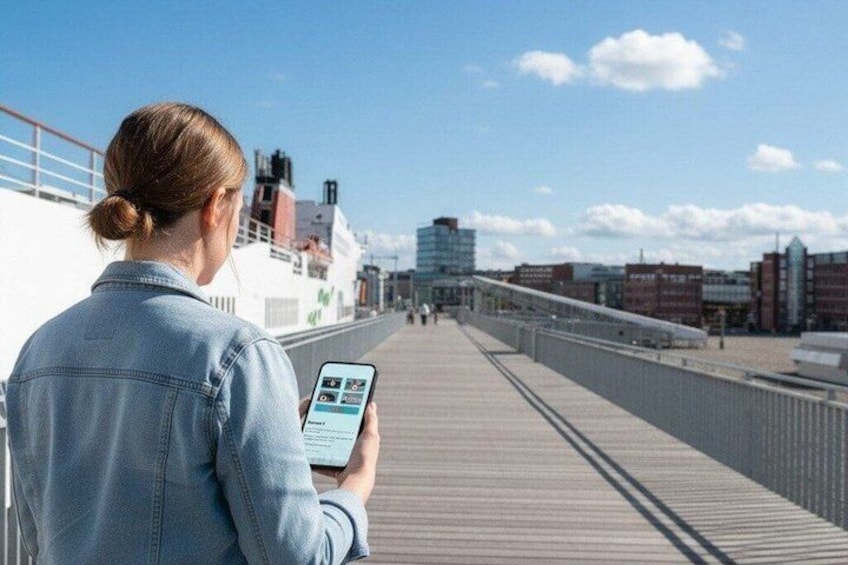 Een persoon op een vogelwachterspad, met een smartphone, kijkt uit over een havenfront met gebouwen en een schip op de achtergrond. 1