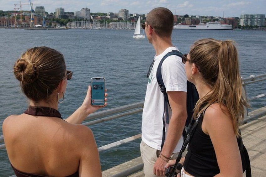 A waterfront scene with a sailboat, people taking photos, and a cityscape in the background. 3