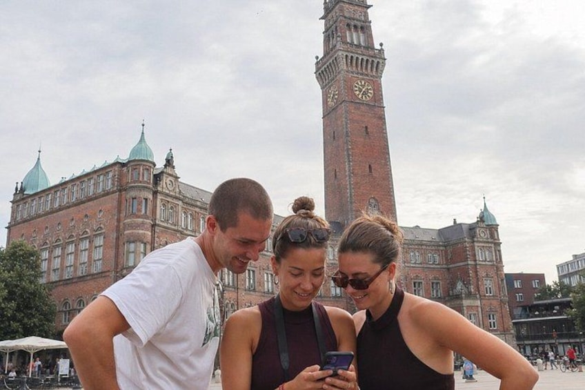 Three people looking at a smartphone in a public square with a historic building and a clock tower in the background. 2