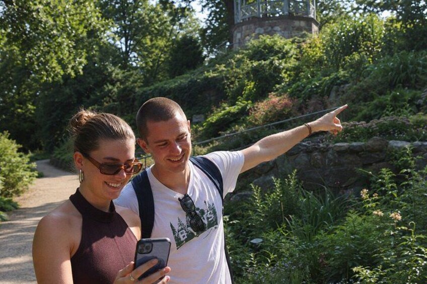 Two people taking a selfie in a park with greenery and a stone structure in the background. 4