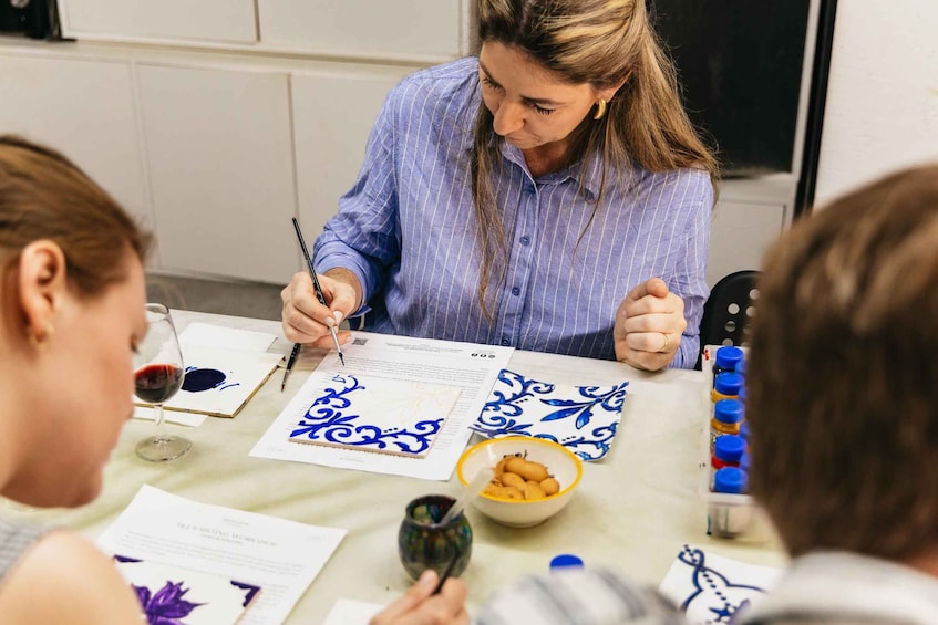 Iemand schrijft met een pen op papier, omringd door blauw-witte papierpatronen, een schaal met lekkernijen en een wijnglas. 1