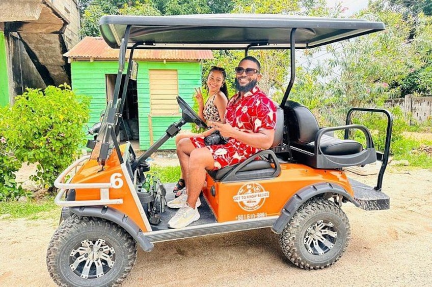 A golf cart with two people, a green building, and a dirt path. 3