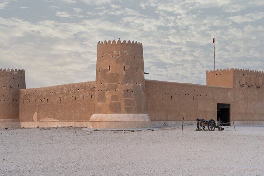Una gran estructura fortificada con una torre central y varias torres más pequeñas, rodeada por una explanada arenosa con una bandera en la cima. 1
