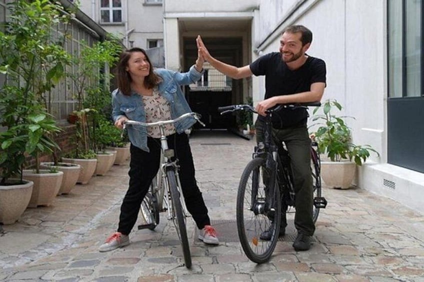 Two people on bicycles in a courtyard with potted plants and a building in the background. 4
