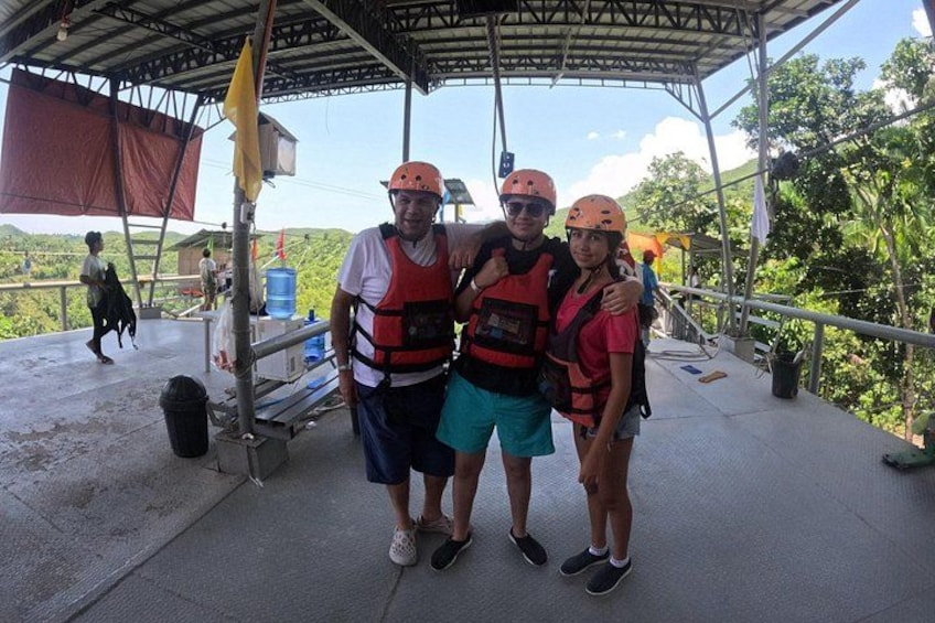 Three people wearing life jackets and helmets standing on a covered platform with a view of greenery and hills in the background. 3
