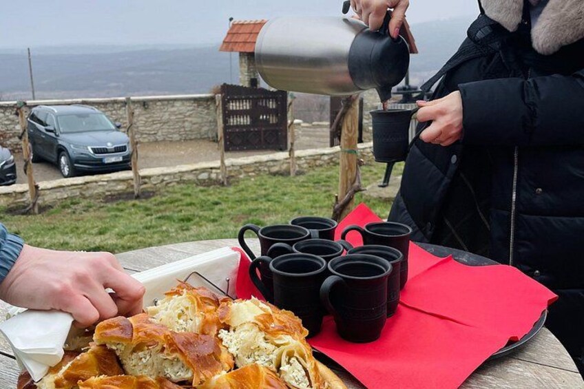 Una persona sirviendo café en una taza, una hogaza de pan y varias tazas negras sobre una mesa con un mantel individual rojo. 2