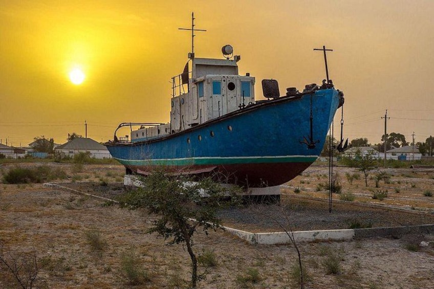 Una barca blu con cabina bianca, ancorata su una riva erbosa e asciutta, con un tramonto sullo sfondo. 1