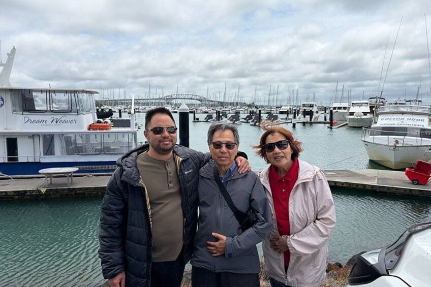 Tres personas junto a un puerto deportivo con barcos y un puente al fondo. 2