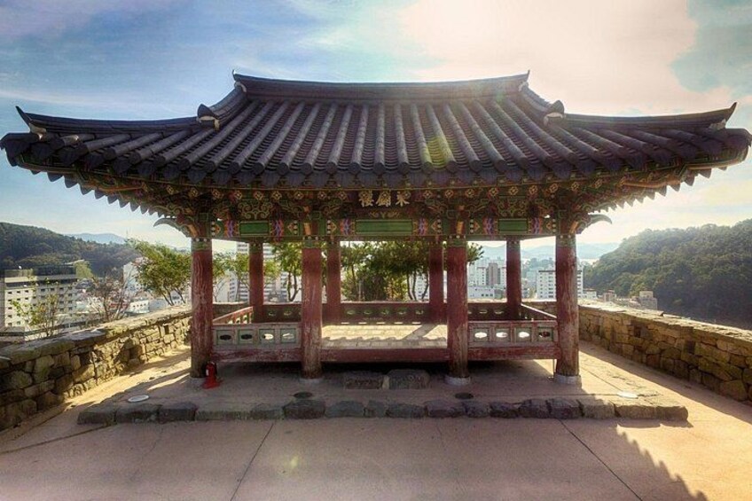 Un padiglione coreano tradizionale con tetto in tegole e decorazioni elaborate, posto su una piattaforma in pietra con vista su una città e delle colline sullo sfondo. 3