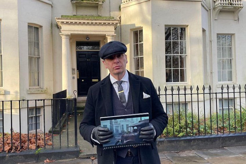 A man in a suit holding a book in front of a building with a black door and a white fence. 1