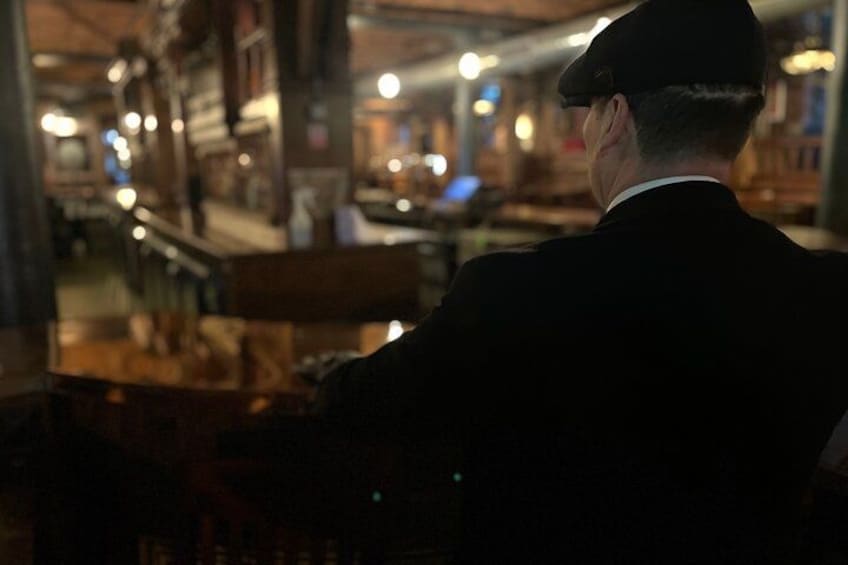 Una persona con traje y sombrero sentada a una mesa en un espacio interior con muebles de madera e iluminación cálida. 5