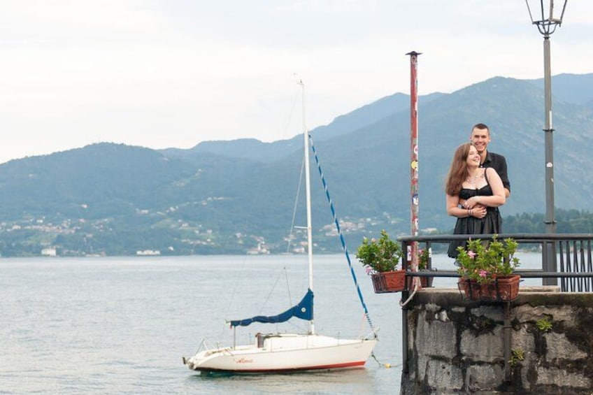 A couple standing by a dock with a sailboat, a stone wall, and mountains in the background. 3