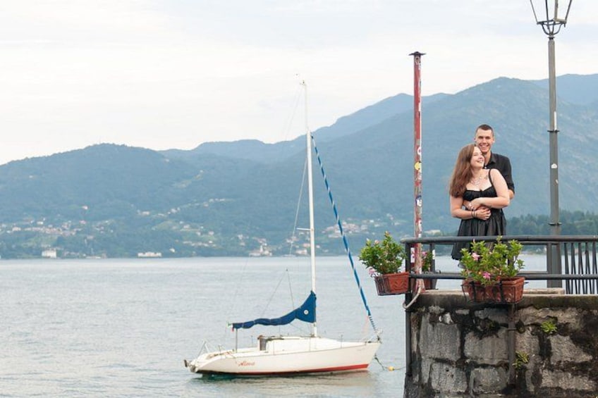 A couple standing by a dock with a sailboat, a stone wall, and mountains in the background. 3