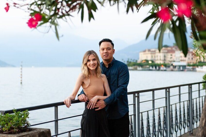 A couple standing by a waterfront with a railing, a body of water, and buildings in the background. 5