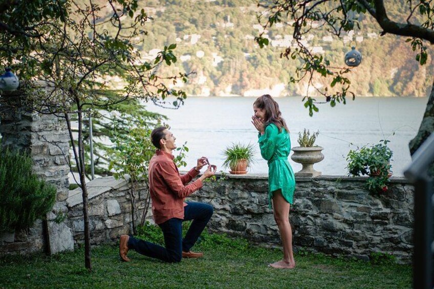 A man proposing to a woman by kneeling on grass with a ring, a stone wall, and a lake in the background. 1