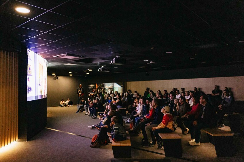 An indoor lecture hall with people seated on benches, facing a presentation screen. 4