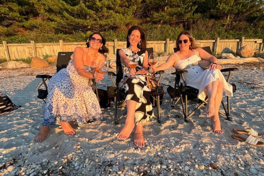 Three people sitting on chairs on a sandy beach with a wooden fence and trees in the background. 4